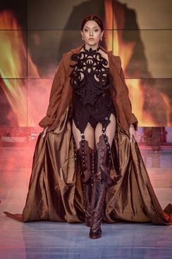 Modelka kroczy po wybiegu w efektownym brązowym płaszczu, pod którym ma na sobie czarny, misternie zaprojektowany body oraz wysokie, sznurowane buty, a za nią rozciąga się ogniste cyfrowe tło.